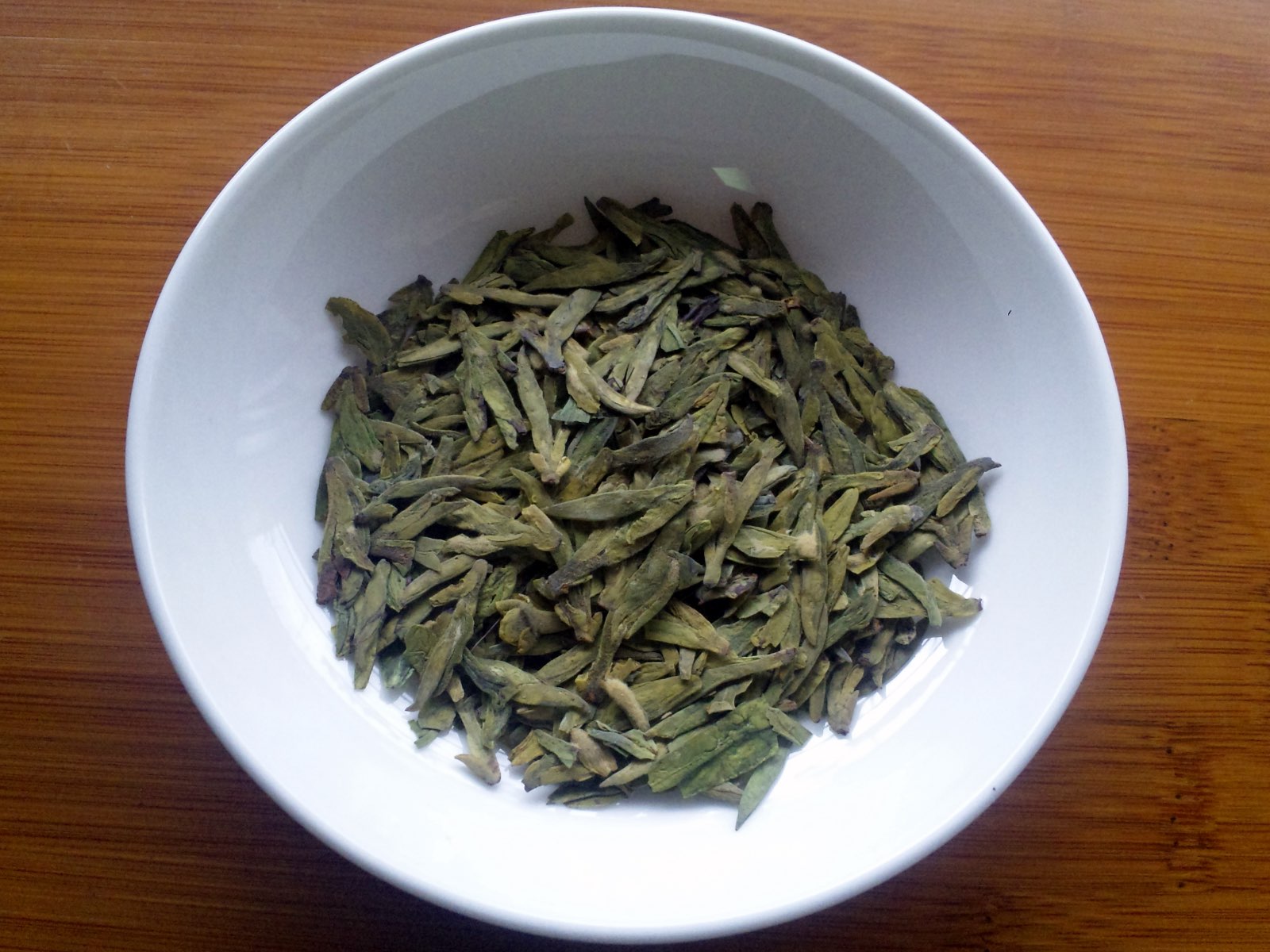 Hojas secas de té longjing, planas y alargadas, de color verde apagado, en un cuenco blanco